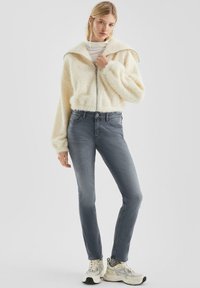Giacca zip con zip di colore crema, morbida e pelosa, abbinata a jeans slim fit grigio scuro e scarpe da ginnastica leggere con accenti a motivo.