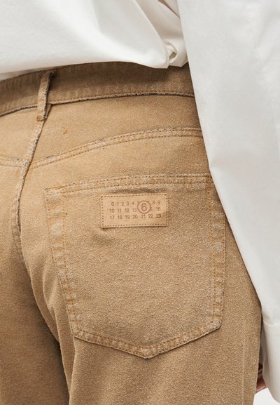Κοντινό πλάνο σε σουέτ παντελόνι καμηλό χρώματος που δείχνει την πίσω τσέπη με κεντημένη ετικέτα με αριθμούς και τον αριθμό 6 σε κύκλο, φορεμένο με λευκό πουκάμισο.