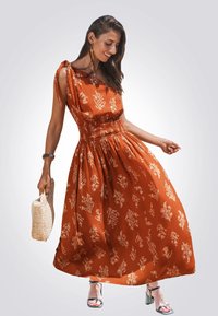 Maxikleid in Rost mit weißem Blumenmuster, gerafftem Bund und Bindeschultern. Kombiniert mit einer geflochtenen Tasche und Sandalen mit transparenten Absätzen.