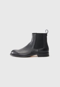Botín Chelsea de cuero negro con parte superior texturizada, paneles laterales elásticos y una etiqueta de tirón. Presenta una suela de goma plana y un diseño de punta redondeada.