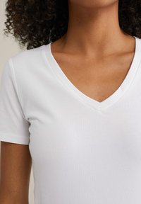 Femme aux cheveux bouclés portant un t-shirt blanc uni à col en V et à manches courtes, vue du cou jusqu'au haut du torse.