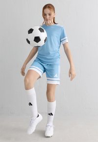 Joven con uniforme de fútbol azul claro haciendo malabares con un balón de fútbol blanco y negro en interiores contra una pared blanca.