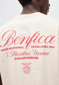 Άνδρας που φοράει μπεζ πουκάμισο με κόκκινο κείμενο "Μπενφίκα", "Μοναδικός και αιώνιος", "Ιδρύθηκε το 1904", "E Pluribus Unum", "85 μεγάλοι τιμητικοί τίτλοι" και ένα κόκκινο έμβλημα.