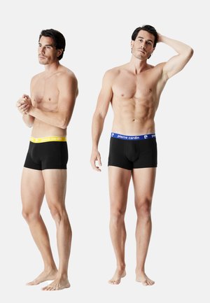 Caleçons boxers noirs avec des ceintures contrastantes colorées, l'une jaune et l'autre bleue, sur deux figures masculines, montrées de la taille vers le haut, pieds nus.