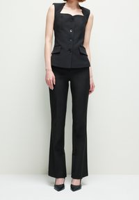 Femme portant un blazer noir ajusté sans manches avec des boutons, un pantalon noir droit assorti et des talons noirs à bout pointu, debout devant un fond blanc.