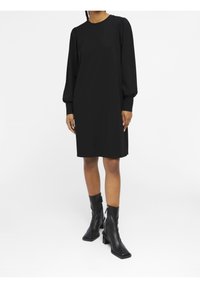 Schwarzes langärmliges Kleid mit lockerer Passform und rundem Halsausschnitt, kombiniert mit knielangen schwarzen Lederstiefeln mit Absätzen und Reißverschlüssen.