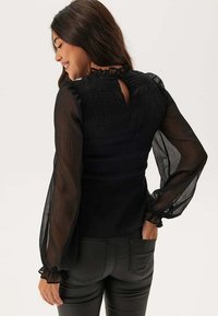 Blusa nera con maniche a sbuffo trasparenti, corpino leggermente strutturato, scollatura a volant e dettaglio a chiave sulla schiena. Abbinata a pantaloni neri attillati.