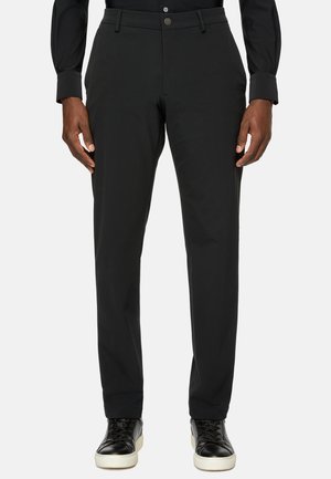 Pantalon noir en tissu lisse et extensible. Présente un devant plat, des passants de ceinture et une coupe ajustée. Associé à des baskets noires.