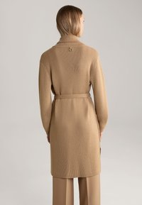 Langer, gerippter Strickcardigan in Hellbraun, mit einem taillierten Gürtel, einem hohen Kragen und einem kleinen Akzent am hinteren Halsausschnitt.