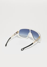 Occhiali da sole bianchi con lenti blu, forma angolare, dettagli in oro sui templi e punte nere sulle aste.