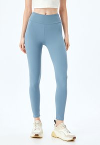 Leggings de color azul claro con una textura suave, cinturilla alta y diseño ajustado, combinados con zapatillas deportivas blancas y beige.