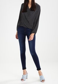 Blusa nera con pois bianchi, jeans scuri a vita alta e sandali con tacco annodati di colore azzurro chiaro. Tessuto morbido, design aderente, maniche arrotolate.