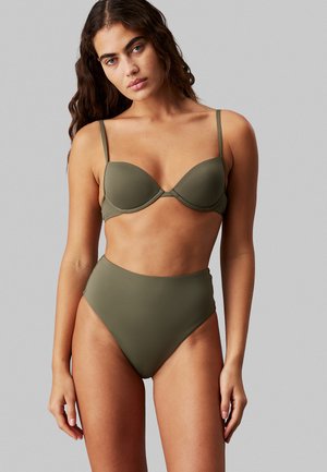 Vrouw met lang golvend haar, gekleed in een olijfgroene, hooggetailleerde bikinibroek en bijpassende onderwired bikinibovenstuk tegen een effen grijze achtergrond.