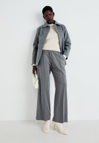 Graue Wollmischung-Jacke, cremefarbener Ripp-Strickpullover, graue weit geschnittene Hose, weiße High-Heel-Stiefel. Kleine weiße Handtasche akzentuiert das Outfit.