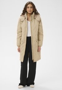 Abrigo largo beige acolchado con capucha, con cremallera frontal completa, bolsillos laterales y puños elásticos. Llevado sobre una camiseta blanca y pantalones negros.