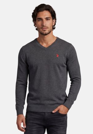 Mann trägt einen dunkelgrauen V-Ausschnitt-Pullover mit einem kleinen roten Logo auf der linken Brust, eine Hand in der Tasche, steht vor einem einfarbigen Hintergrund.