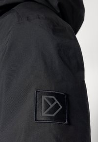 Črn material z gladko teksturo, vsebuje všito kvadratno nalepko z geometrijskim logotipom v temnejši senci črne.