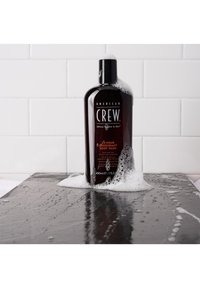 Bouteille de gel douche en plastique marron foncé sur une surface noire, affichant une mousse blanche. L'étiquette comprend "American Crew" et des détails sur le produit en texte blanc.
