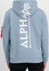 Svijetloplava hoodie napravljena od mekog materijala, s velikim bijelim logom na leđima i malim dodatnim džepom na rukavu.