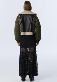 Olivgrüne Steppjacke mit schwarzem Leder-Rücken, beigem Lammfellkragen; getragen über einem schwarzen Maxikleid aus Spitze mit geschichteter Textur.