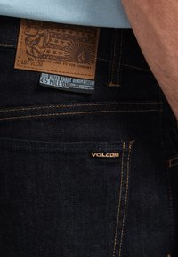 Ciemne dżinsy z denimu mają skórzaną naszywkę z tłoczonym wzorem, żółtym przeszyciem i małym logo marki nad tylną kieszenią.