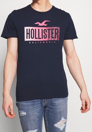 Mężczyzna w granatowej koszulce Hollister California z różowym logo i znoszonych niebieskich dżinsach na jednolitym jasnym tle.