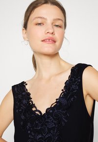 Top preto sem mangas com um decote profundo em V, apresentando um intrincado bordado floral azul-marinho e um acabamento de tecido texturizado.