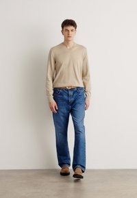 Beige színű, hosszú ujjú V nyakú pulóver kék farmernadrággal párosítva. A farmer laza szabású, barna lábbelival kiegészítve.