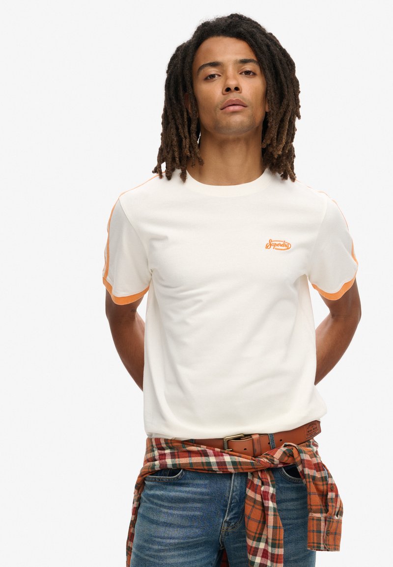Hvid tshirt med korte ærmer og orange detaljer ved skuldrene samt et lille orange logo. Bæres med blå jeans og et rutet bælte knyttet om taljen.