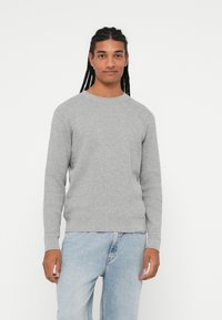 Pulover gri cu pliuri, cu gât rotund și mâneci lungi, asociat cu blugi de denim de un albastru deschis. Textură moale și design ajustat.