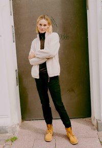 Mudel, kes on riietatud beeži fliisjakki tumeda särgi, mustade laevukottpükste ja beežide saapadega, seistes metallseina taustal.