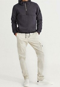 Sudadera con capucha gris oscuro con media cremallera y dobladillo elástico, combinada con pantalones cargo beige claro con bolsillos laterales y cordón. Zapatillas blancas.