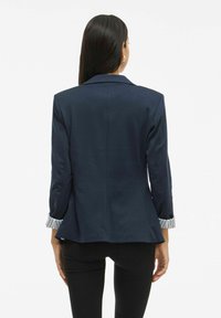 Femeie cu părul lung și închis la culoare, purtând un sacou bleu marin cu manșete în dungi rulate și pantaloni negri ajustați, stând cu spatele.