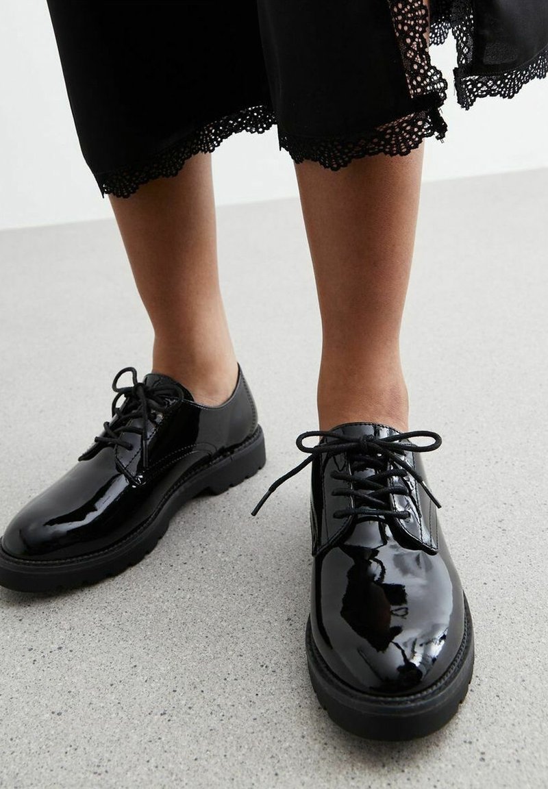 Chaussures à lacets noires brillantes portées avec un pantalon noir court ornée de dentelle, se tenant sur un sol gris clair et moucheté.