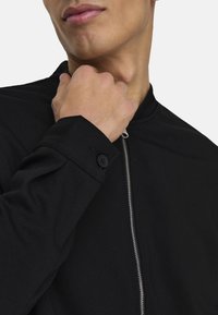 Svart zip-up jakke laget av glatt stoff, med stående krage og knappete mansjett. Nærbilde av hånd nær nakken.