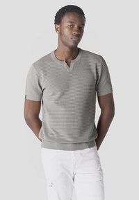 Jonge man met kort krullend haar, gekleed in een lichtgrijs gebreid shirt met korte mouwen en witte gescheurde broek, staand met handen achter de rug.