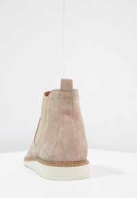 Beige semišové Chelsea boty s elastickými bočními panely a bílou gumovou podrážkou, zobrazené z zadní strany na bílé ploše na jednoduchém bílém pozadí.