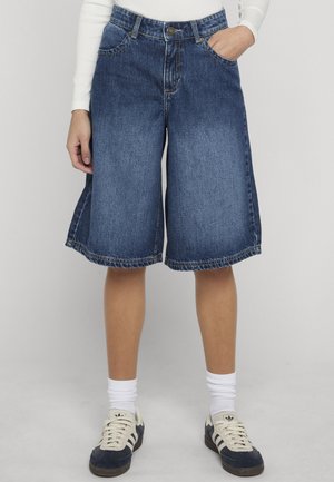 Inimene, kes kannab keskmise pikkusega siniseid teksapükse, valgeid sokke ja must-valgeid tossusid, seisab ühe käe taskus.