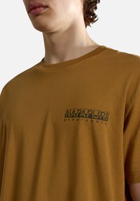 Camiseta de algodón marrón con un logo impreso en negro en el lado izquierdo del pecho. Cuello redondo, mangas cortas y textura suave.