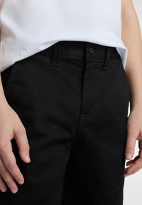 Schwarze Jeans mit klassischem Schnitt, sichtbaren Nähten und einem Frontknopfverschluss. Getragen mit einem weißen kurzärmligen Hemd.