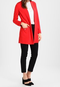 Manteau rouge à manches longues avec une coupe ajustée, devant ouvert et deux poches latérales. Porté sur une chemise blanche et un pantalon noir avec des chaussures noires à talons.