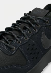 Baskets Nike noires avec lacets épais, tissu texturé, panneau latéral perforé et détails cousus visibles sur un fond clair.