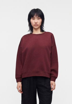 Ung person med glat sort hår iført en løs bordeaux sweatshirt og sorte bukser, stående foran en ensfarvet hvid baggrund.