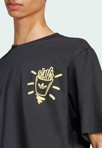 Maglietta a maniche corte in cotone nero con una grafica di una tazza di patatine fritte con il logo Adidas, evidenziata da accentuazioni giallo chiaro. Vestibilità rilassata.
