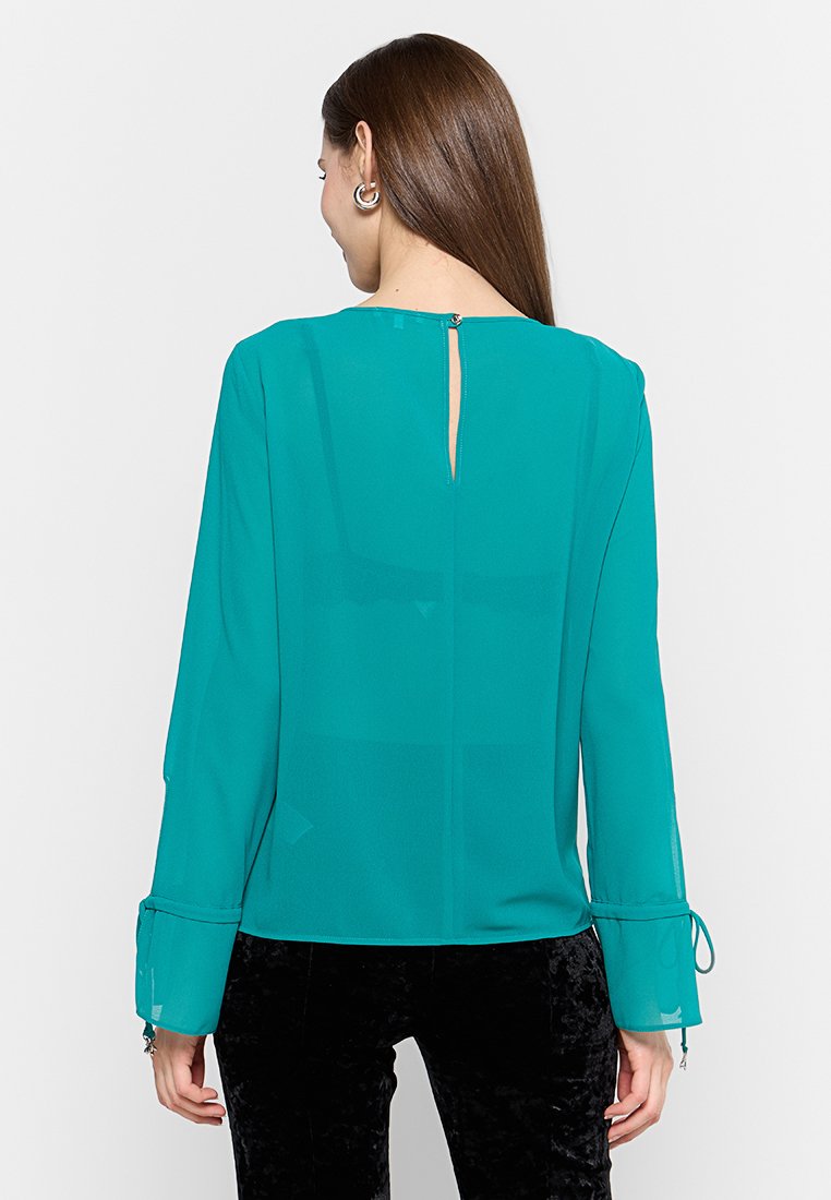 Türkise Bluse aus leichtem, transparentem Stoff, mit langen ärmeln und Bindebändern, einem Ausschnitt im Rücken und einem geraden Saum. Kombiniert mit schwarzen Hosen.