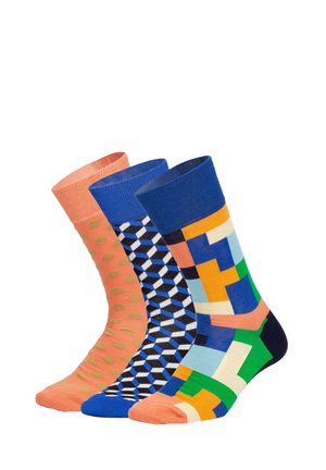 Trzy kolorowe skarpetki z wzorami: pomarańczowa z złotymi kropkami, niebieska z trójwymiarowymi kostkami oraz wielokolorowa z geometrycznymi kształtami, stojące obok siebie.