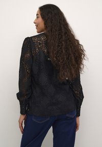 Blusa in pizzo nero con motivi floreali, maniche lunghe, polsini con bottone e vestibilità rilassata. Tessuto strutturato con sovrapposizione trasparente.
