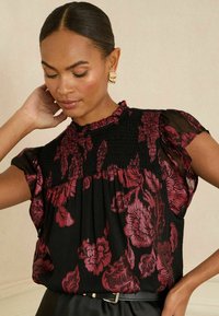 Juoda bluzelė su raudonais gėlių raštais, smulkintais rankogaliais ir trumpomis permatomomis rankovėmis. Medžiaga turi blizgų paviršių ir laisvą pjūvį.