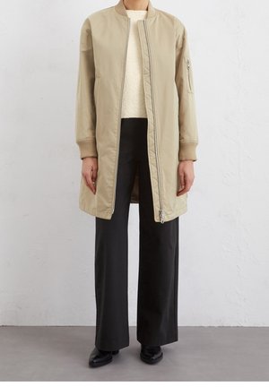 Person trägt eine beige lange Bomberjacke, cremefarbenen Pullover, schwarze weite Hosen und schwarze Schuhe und steht vor einer weißen Wand.