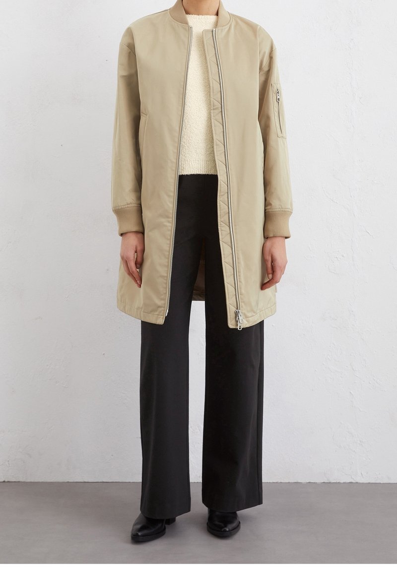 Personne portant une longue veste bomber beige, un pull crème, un pantalon noir à jambes larges et des chaussures noires, debout contre un mur blanc.